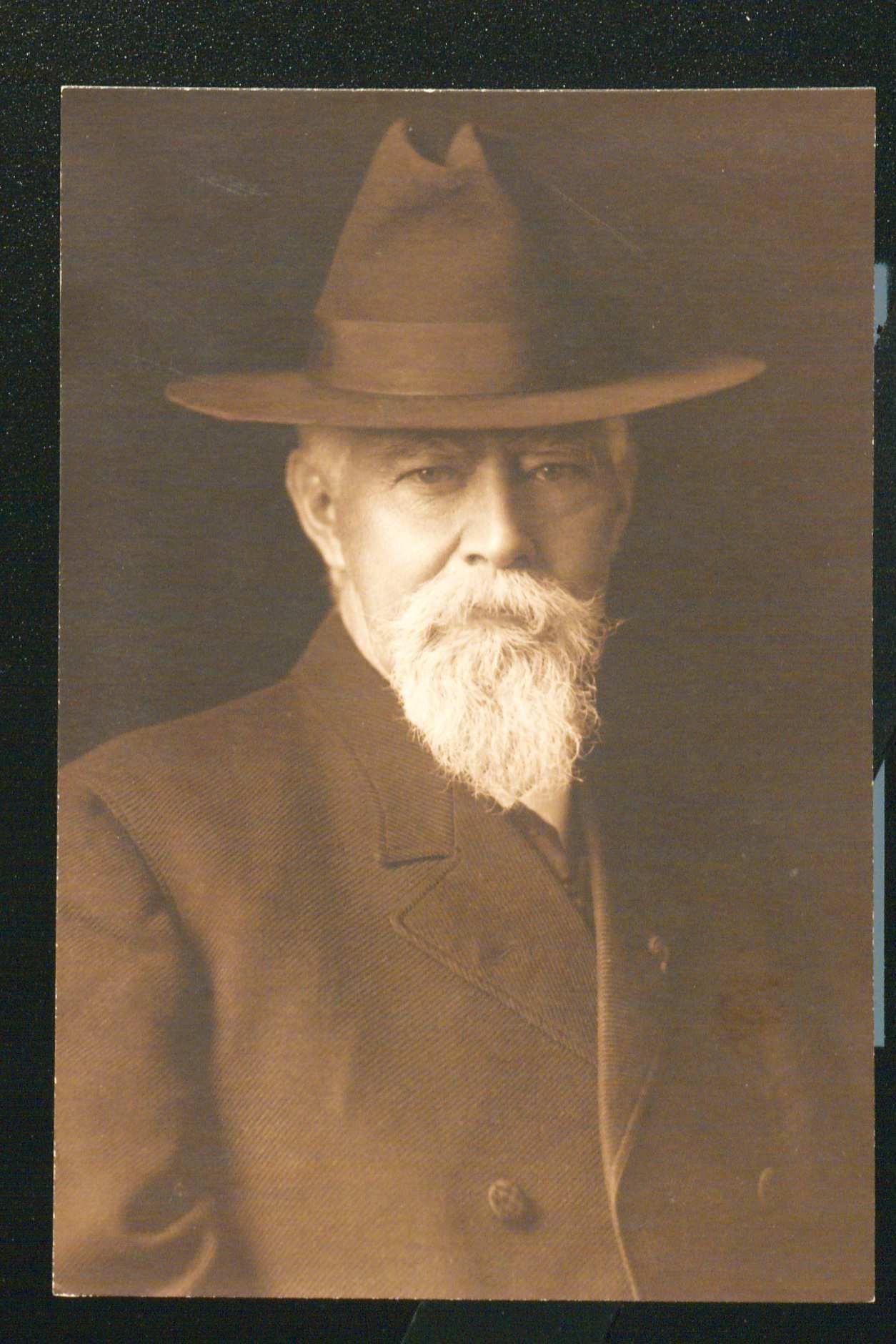 Portrait of John Black looking straight into the camera. He is wearing a wide brimmed hat with a white moustache and beard.He is wearing an overcoat