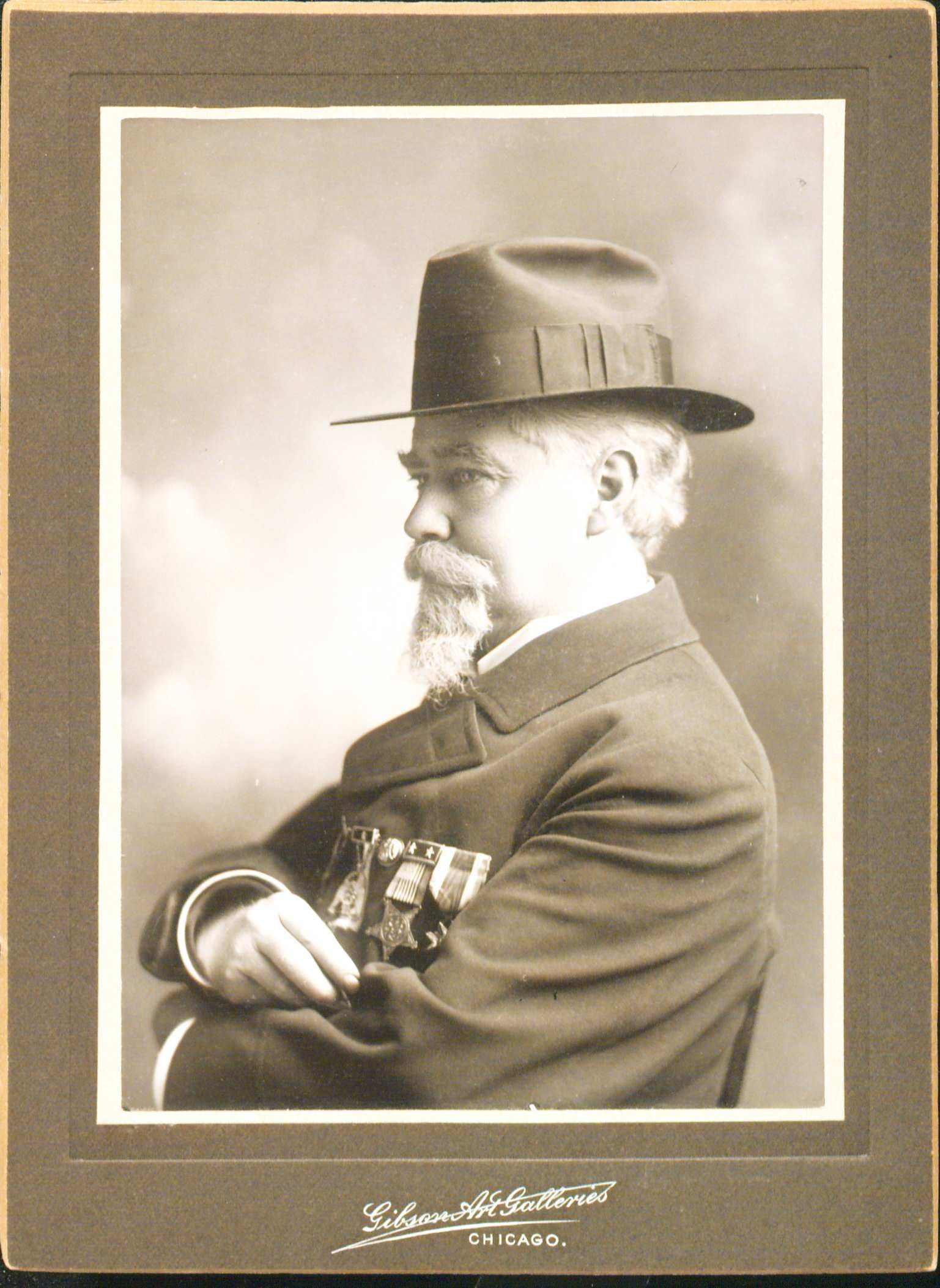 Portrait of John Black sitting down with his arms crossed facing the left. He is wearing a hat an overcoat and there are medals hanging from his left breast pocket.