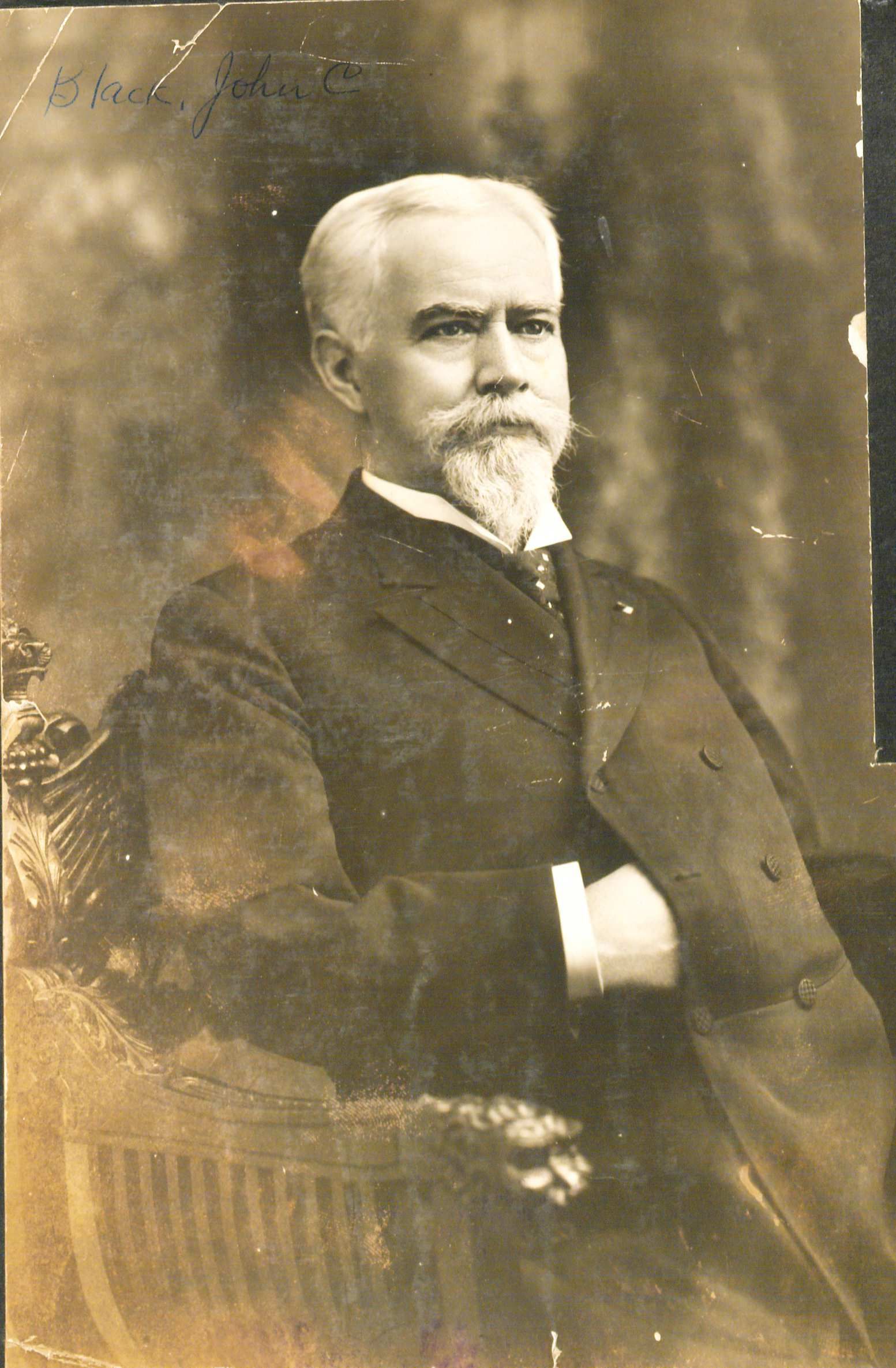 Portrait of John Black sitting down looking off to the right. He has white hair and a moustache and a beard.