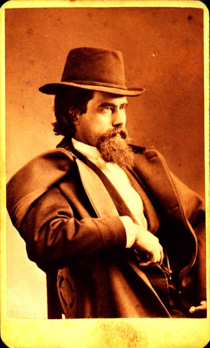 Portrait of John Black sitting down in a chair facing off to the right. He is wearing a hat and an overcoat over his suit