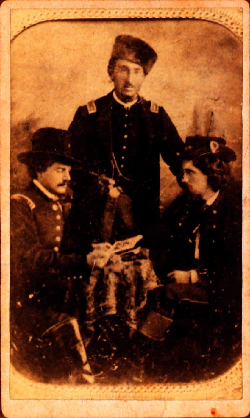 PIcture of 3 men. Two are sitting and facing each other. The third man is standing in the middle facing forward. All are wearing military uniform