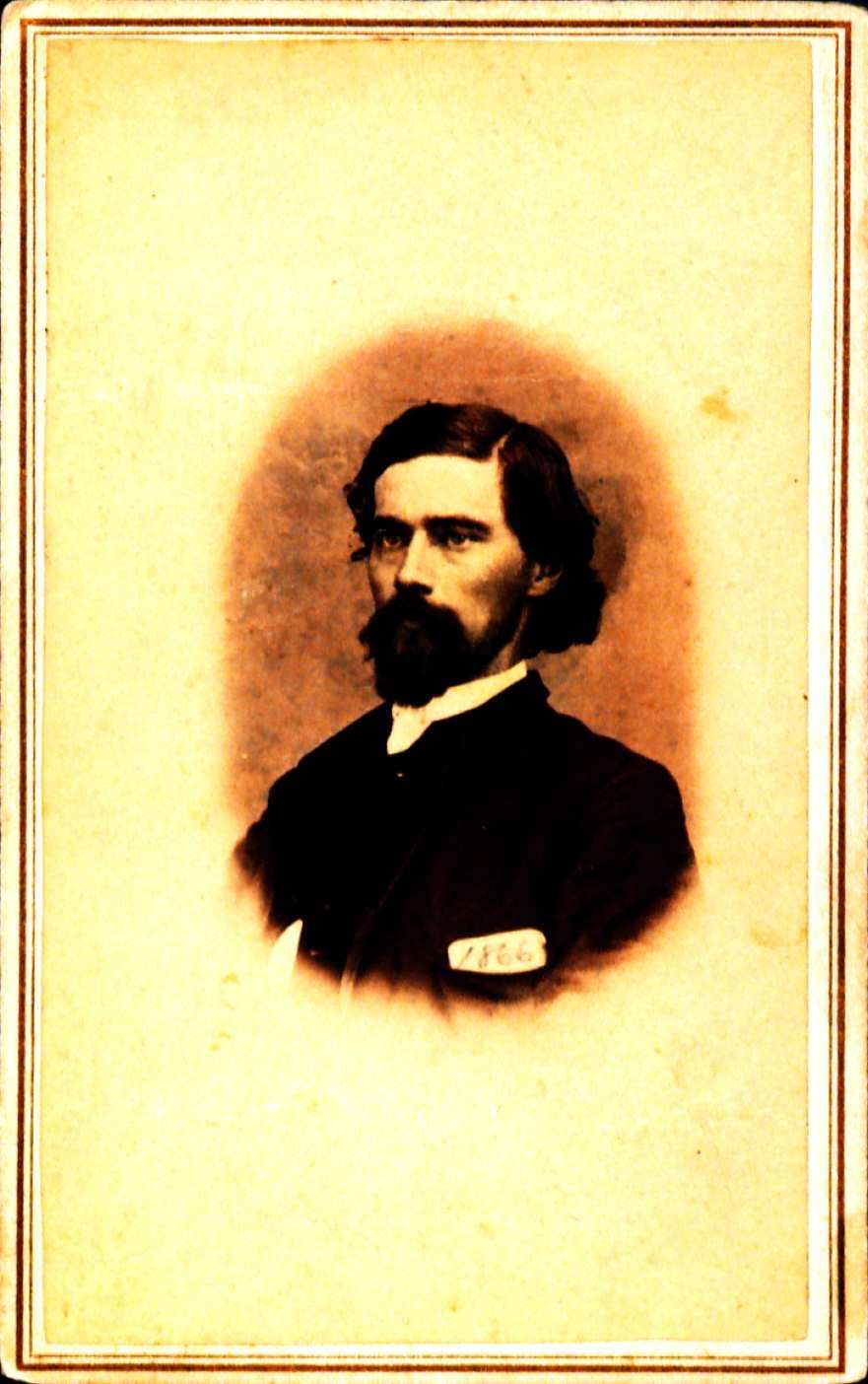 Portrait of John Black looking off to the left. He has a beard is wearing a whtie shirt and a black suit