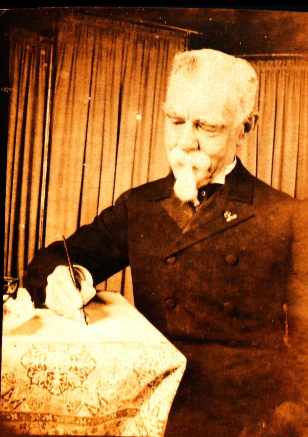 Picture of John Black holding a pen and looking down at what he's writing. he has short white hair a white oustache and a white beard. He is wearing a dark colored suit