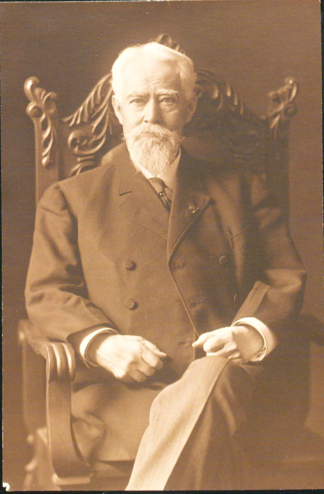 Portrait of John Charles Black sitting in a tall backed chair. He has a short white hair with a white moustache and beard. He is wearing a suit with an overcoat