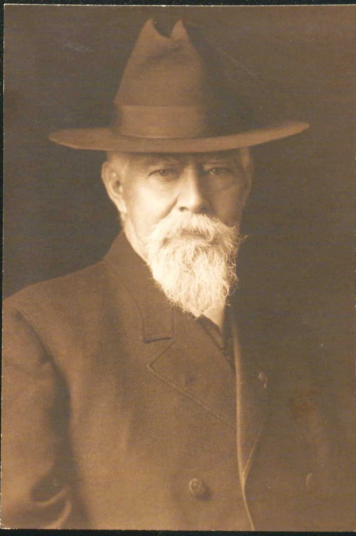 PIcture of John Charles Black looking off to the left. He is wearing a black hat and wearing double breasted jacket. He has a moustache and beard