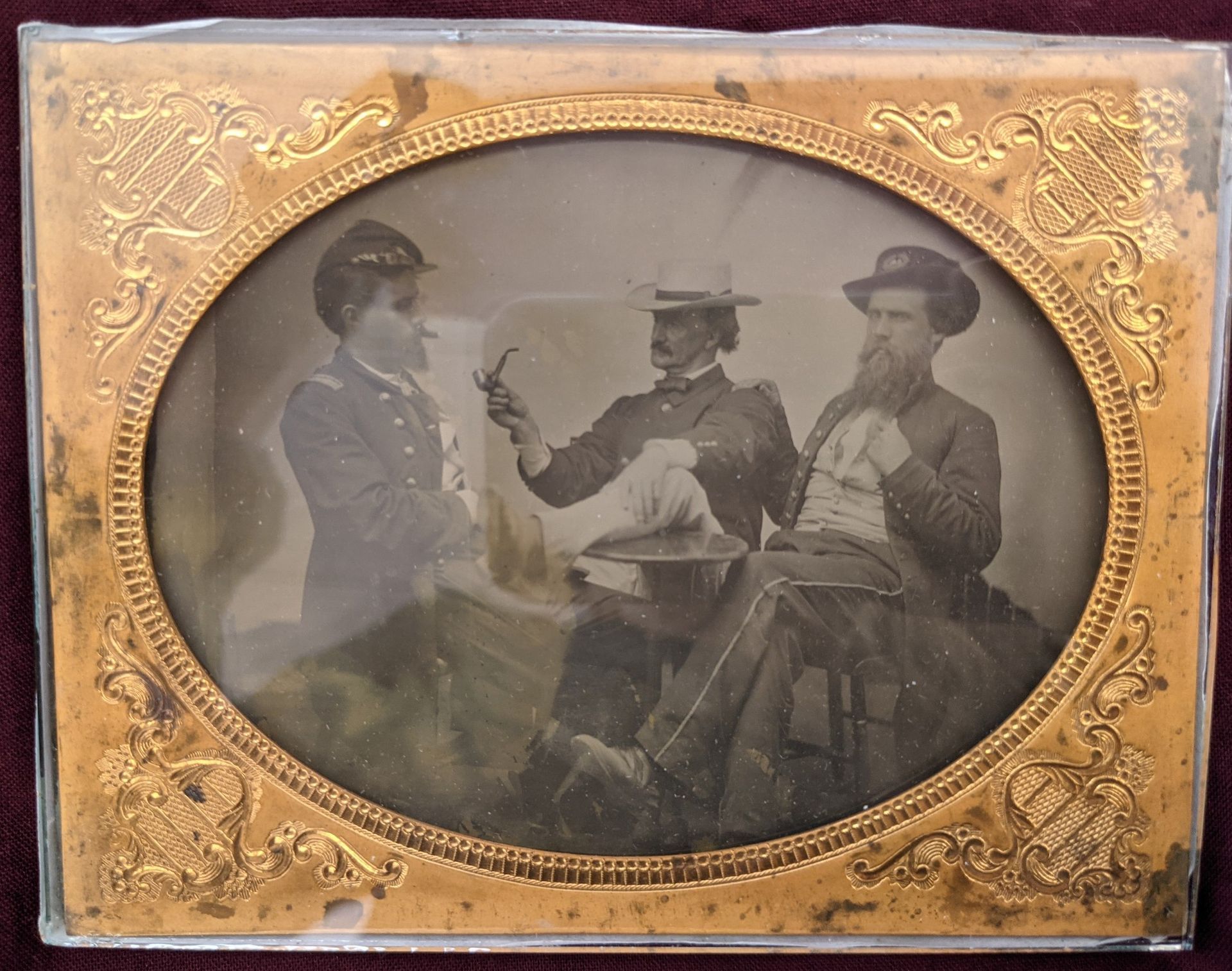 Portrait of John Black facing off to the left. He has a moustache and a beard. He is wearing a white shirt with a whitPicture of 3 men in military unifrom sitting around a table, one is holding a pipe. two are looking off to the side. the picture is in a gold framee bowtie and a black suit jacket