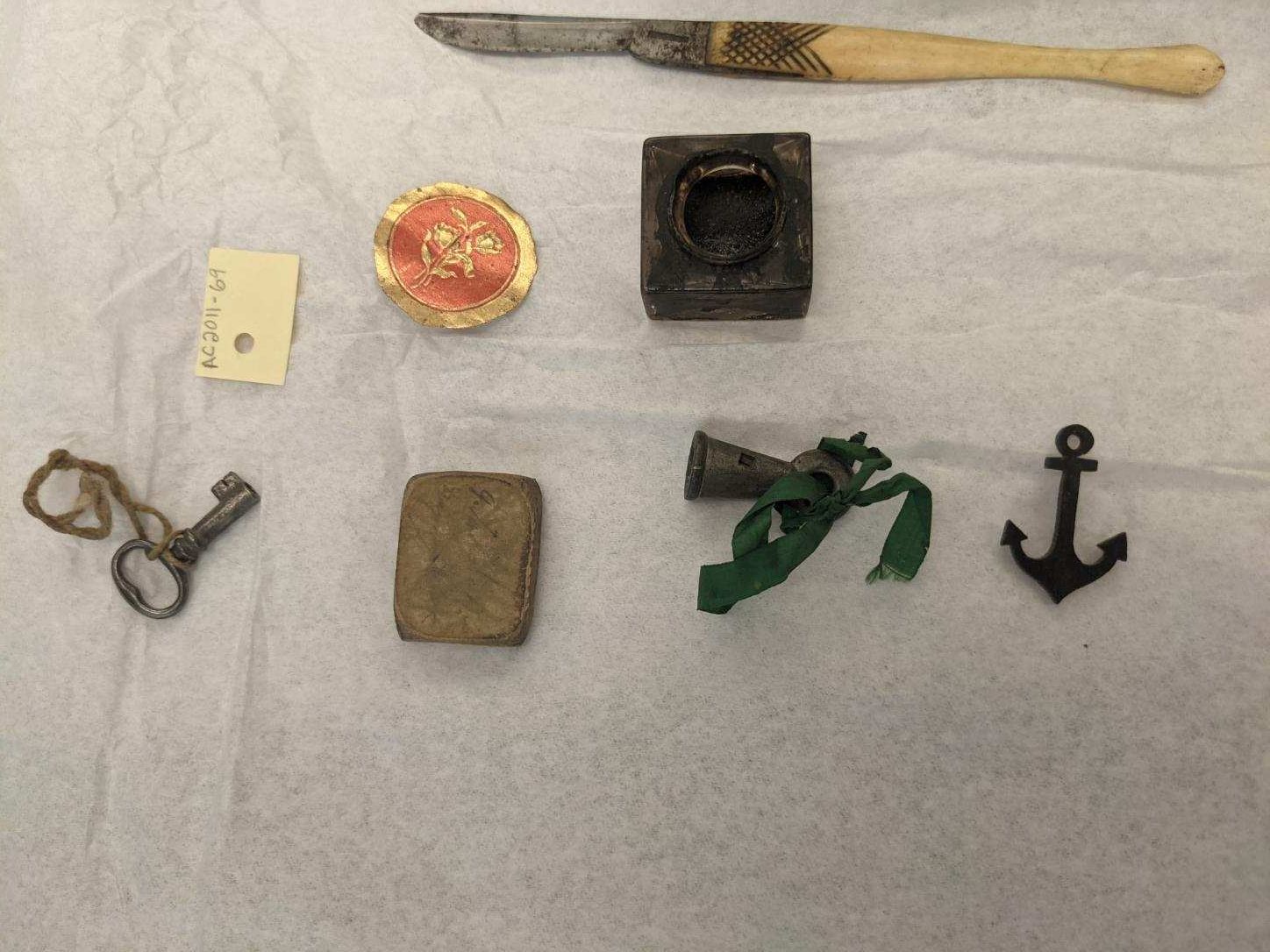 Picture of an assortment of writing utensils. There is a square bottle of ink, a key, the lid for the bottle of ink, a bell with a green sash threaded through the top and a small metal anchor