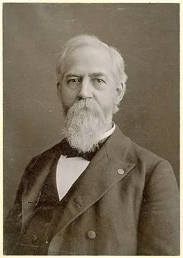 Portrait of William Black looking straight into the camera. He has a moustache and a beard. He is wearing a white shirt, dark colored bow tie, vest and overcoat.