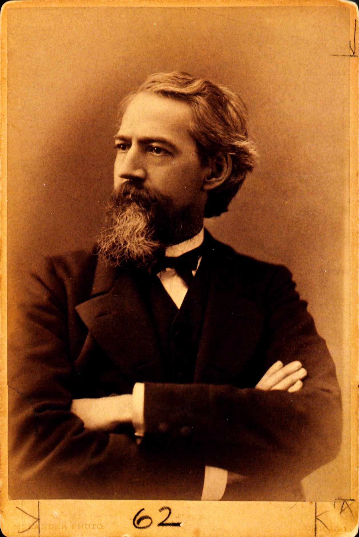William Black with a moustache and beard. Wearing a white shirt and black Suit with a black nec tie. His arms are folded and is looking off to the left.