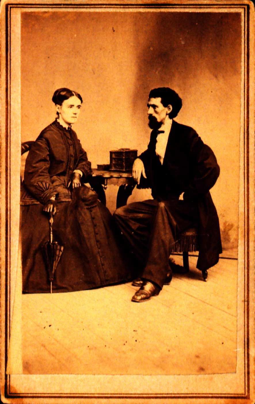 Picture of William Black and a woman both are sitting in a chair. He has his arm propped up on a table between them. She has one arm propped up on the table and one hand holding a folded up umbrella. The umbrella is pointing towards the ground