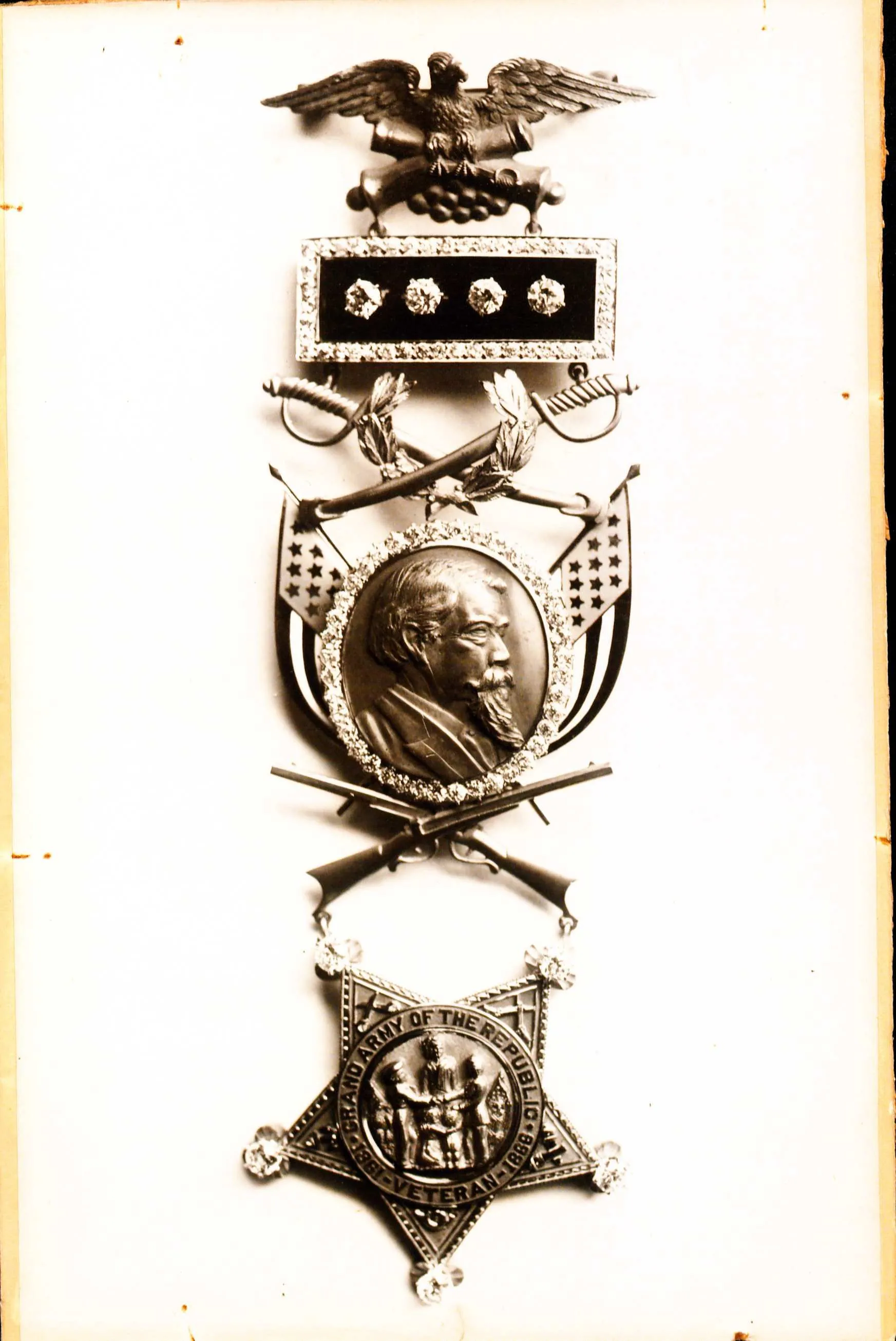 PIcture of a medal of honor. there is a bar at the top with 4 buttons on it, two swords and a shield dangle off the bar. in the middle of the shield is a picture of John Black. Under his portrait are two crossed rifles. Under the rifles hangs a 5 pointed star